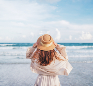 Frau mit Strohhut steht am Strand und blickt aufs Meer, w&auml;hrend der Wind ihr Haar bewegt.