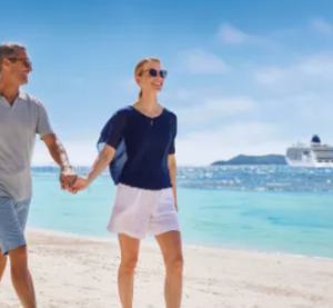 Ein Paar spaziert entspannt am sonnigen Sandstrand entlang, w&auml;hrend im Hintergrund ein Kreuzfahrtschiff von Hapag-Lloyd Cruises im t&uuml;rkisblauen Meer ankert.