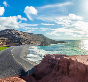 Küstenstraße auf Lanzarote mit schwarzem Lavastrand, rötlichen Felsen und Blick auf den Atlantik unter blauem Himmel.