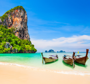 Traditionelle Longtailboote am tropischen Sandstrand mit türkisfarbenem Wasser und Kalksteinfelsen in Thailand bei sonnigem Wetter.