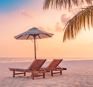 Zwei leere Liegest&uuml;hle unter einem Sonnenschirm am Sandstrand bei Sonnenuntergang, mit Palmen im Vordergrund.