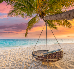 H&auml;ngesessel unter einer Palme an einem tropischen Sandstrand bei farbenfrohem Sonnenuntergang.