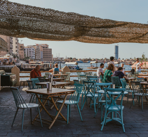 Straßencafé mit türkisfarbenen Stühlen am Dubai Creek, Menschen sitzen im Schatten mit Blick auf Boote und Skyline.