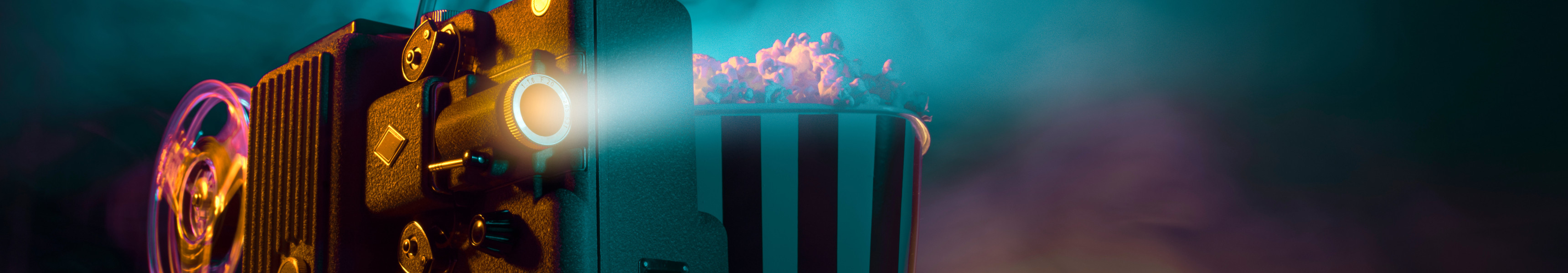 Alter Filmprojektor mit laufender Filmrolle, daneben eine gestreifte Popcornt&uuml;te, im Hintergrund bunte Licht- und Rauch&shy;effekte.