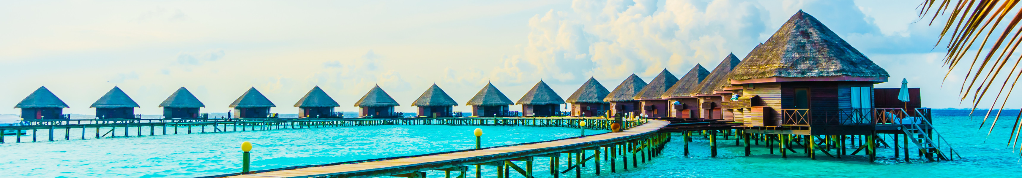 Reihen von Überwasser-Bungalows mit Strohdächern auf den Malediven, verbunden durch Holzstege über türkisblauem Meer, eingerahmt von Palmenblättern im Vordergrund.