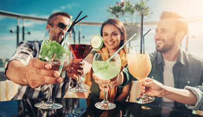 Gruppe junger Leute stößt mit Cocktails auf einer sonnigen Rooftop-Bar an.