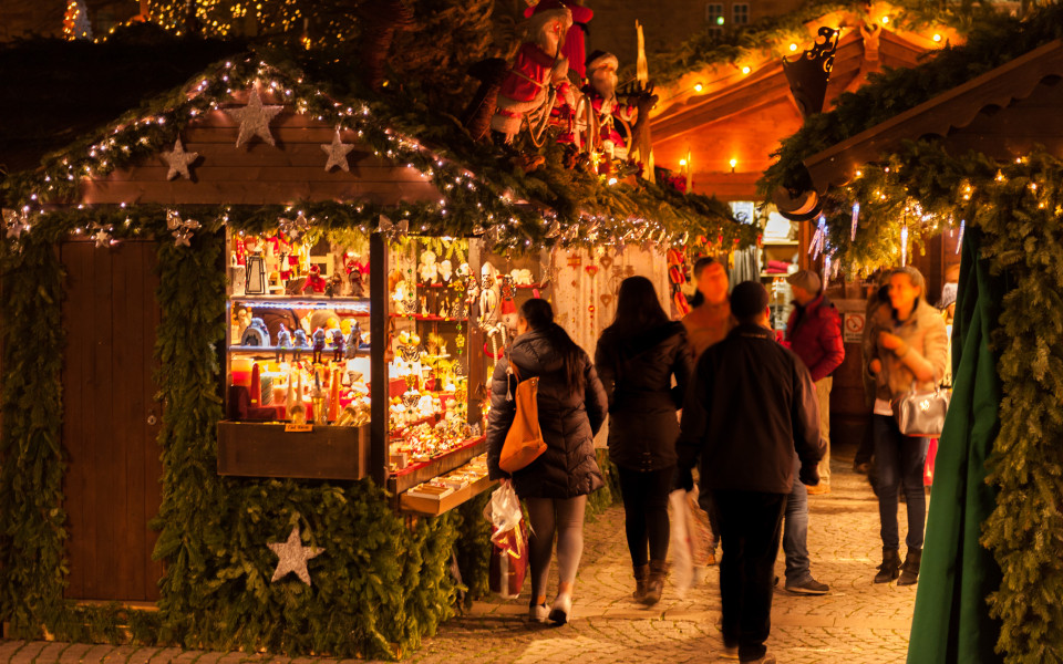 Stimmungsvoll beleuchtete Weihnachtsmarktst&auml;nde in Paris, geschm&uuml;ckt mit Tannenzweigen und Lichterketten, w&auml;hrend Besucher festliche Dekorationen und handgefertigte Geschenke entdecken.