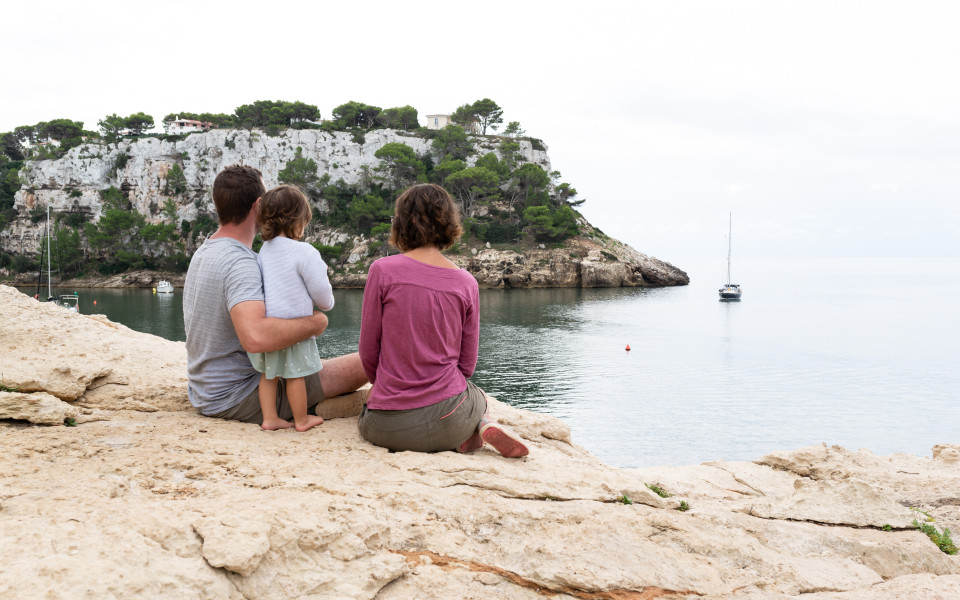 Familie sitzt auf einem Felsen am Wasser in Menorca und genie&szlig;t die Aussicht auf das Meer und die umliegenden Klippen. 