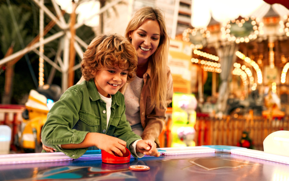 Mutter und Sohn spielen zusammen an einem Airhockey-Tisch im Bayern Park, umgeben von bunten Lichtern und Fahrgeschäften.