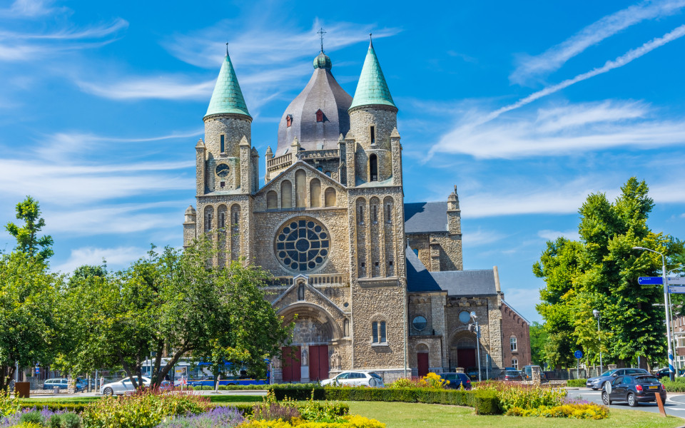 Romanische Kirche in Maastricht mit markanten T&uuml;rmen, gr&uuml;nlichen D&auml;chern und einer gro&szlig;en Rosette, umgeben von B&auml;umen, Blumen und einer gepflegten Parkanlage.